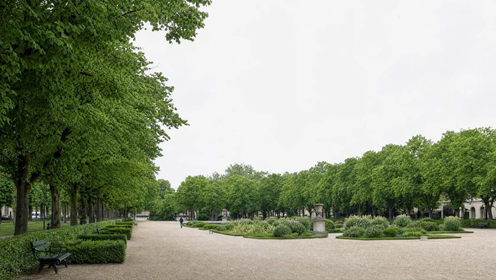 A tree-lined civic square in full leaf with gravel walks and benches, a few people at far distance, nobody identifiable.