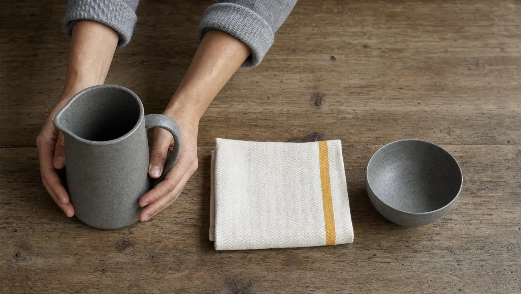 Seen from above: hands setting a jug, a folded tea towel and a small bowl in a row on a scrubbed wooden worktop, nothing labelled.