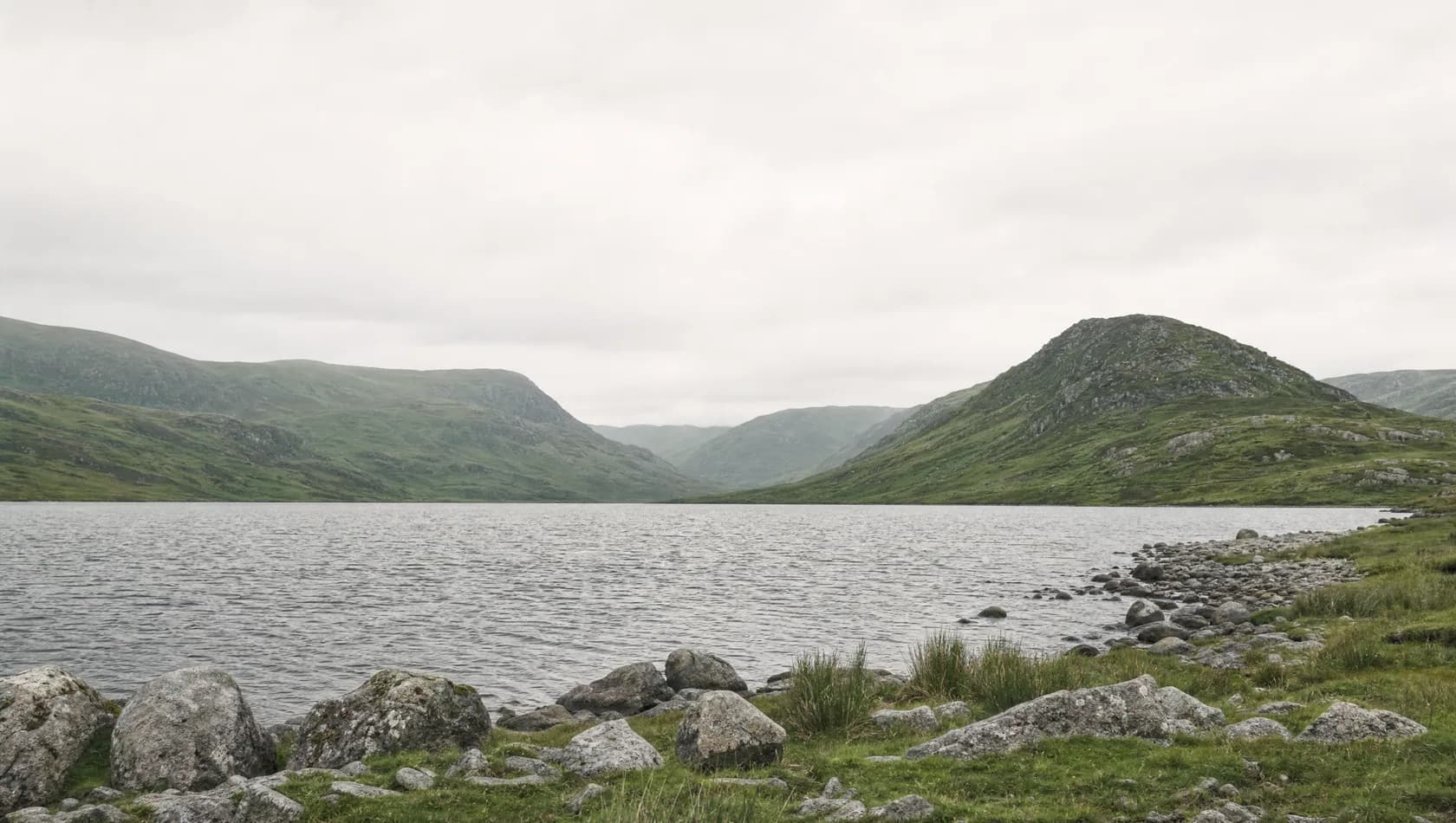 A wide upland lake with a stony foreshore and hills folding away behind it under an even grey sky, no people.