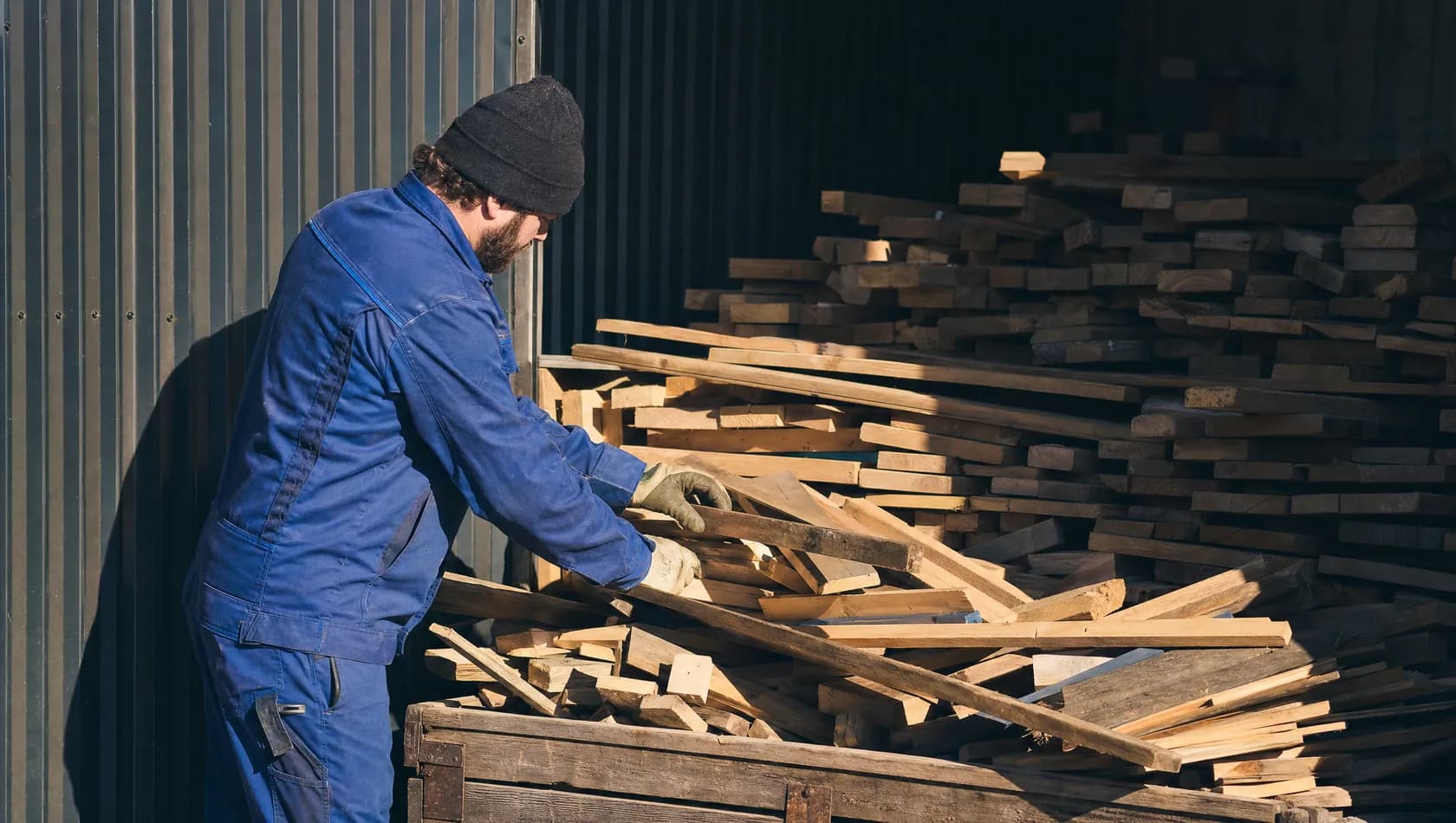 One person sorting timber offcuts in a yard under hard afternoon sun, the shade line cutting sharply across the stack.