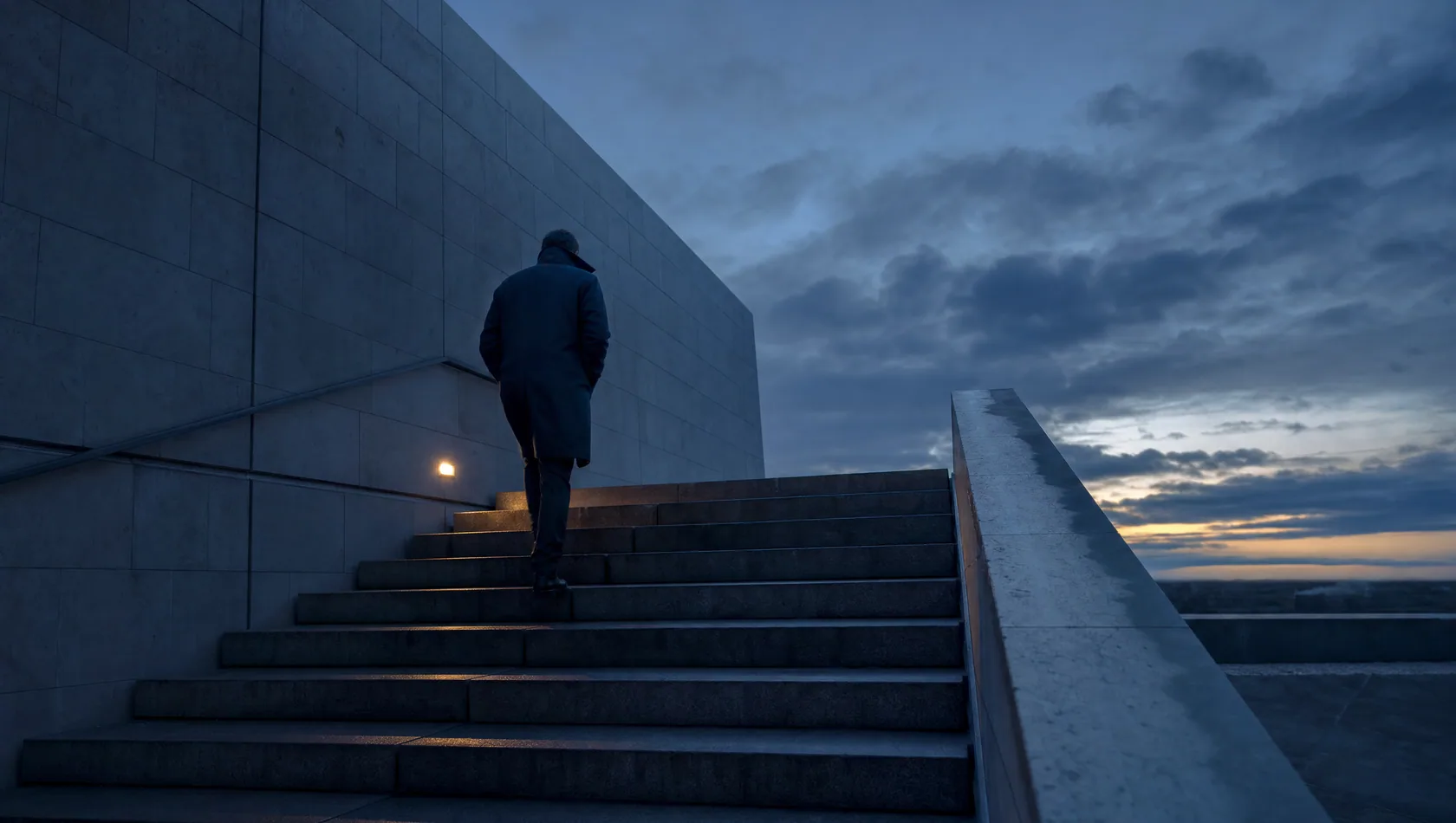 A figure climbing a long flight of civic steps in blue pre-dawn light, coat collar up, seen from below and behind.
