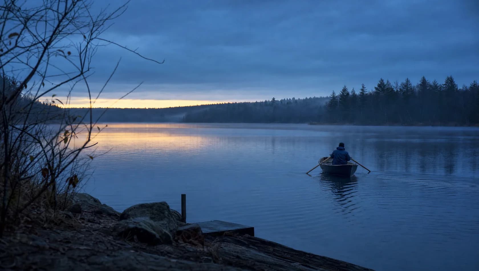 A lone figure rowing a small boat on still water at first light, seen from the bank at distance, cold blue with a thin warm line on the far shore.