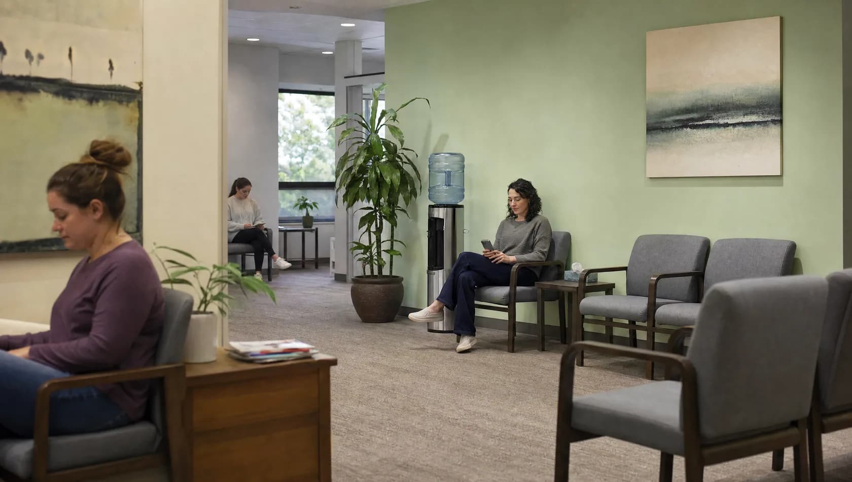 A clinic waiting area at middle distance with chairs, a water cooler and a plant, two people seated apart.