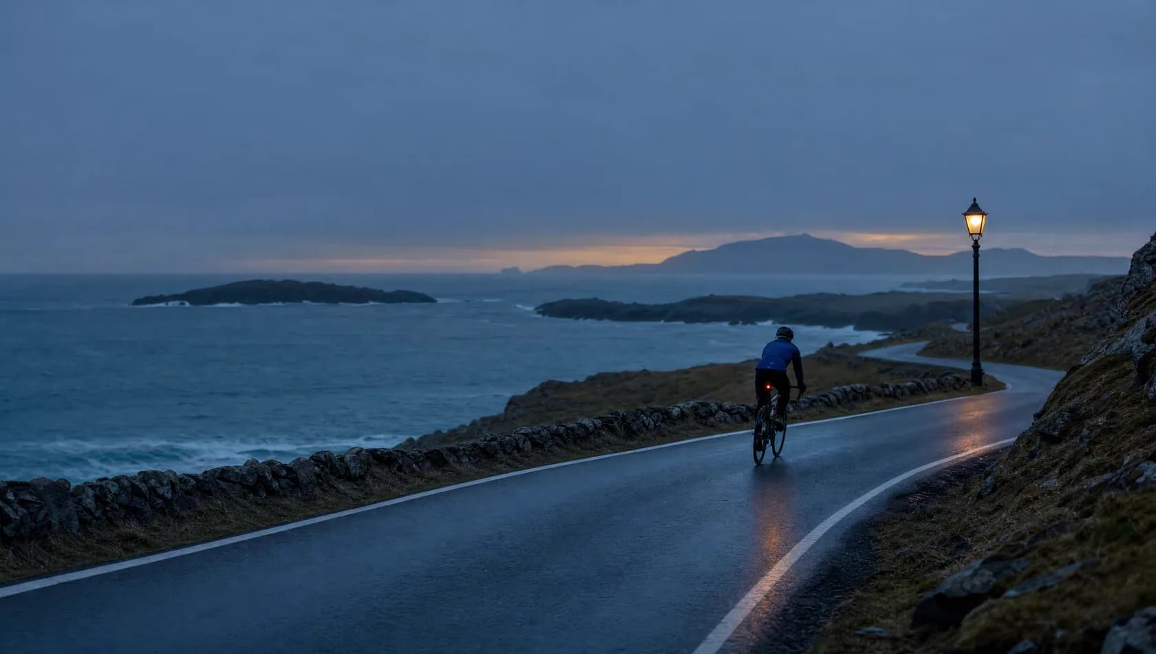 A lone cyclist on an empty coast road at first light, small against a blue-grey sea, one warm lamp still lit on the verge.