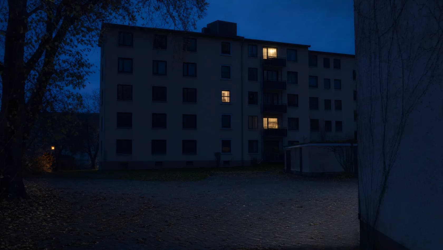A block of flats across a dark courtyard at night with three windows lit at different floors and the rest dark.