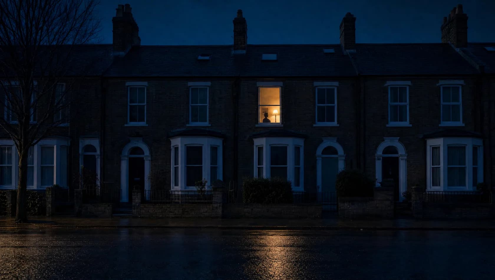A terrace of houses seen from the dark wet street at night with one upstairs window lit and the rest unlit.