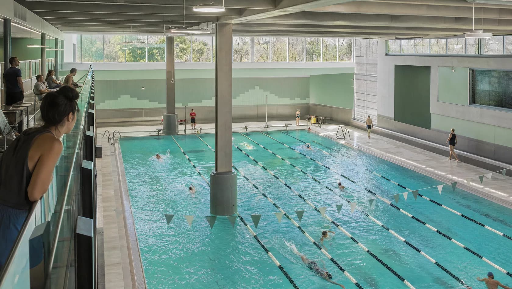 A municipal pool hall seen from the balcony with a handful of swimmers in the lanes, daylight through clerestory glass.