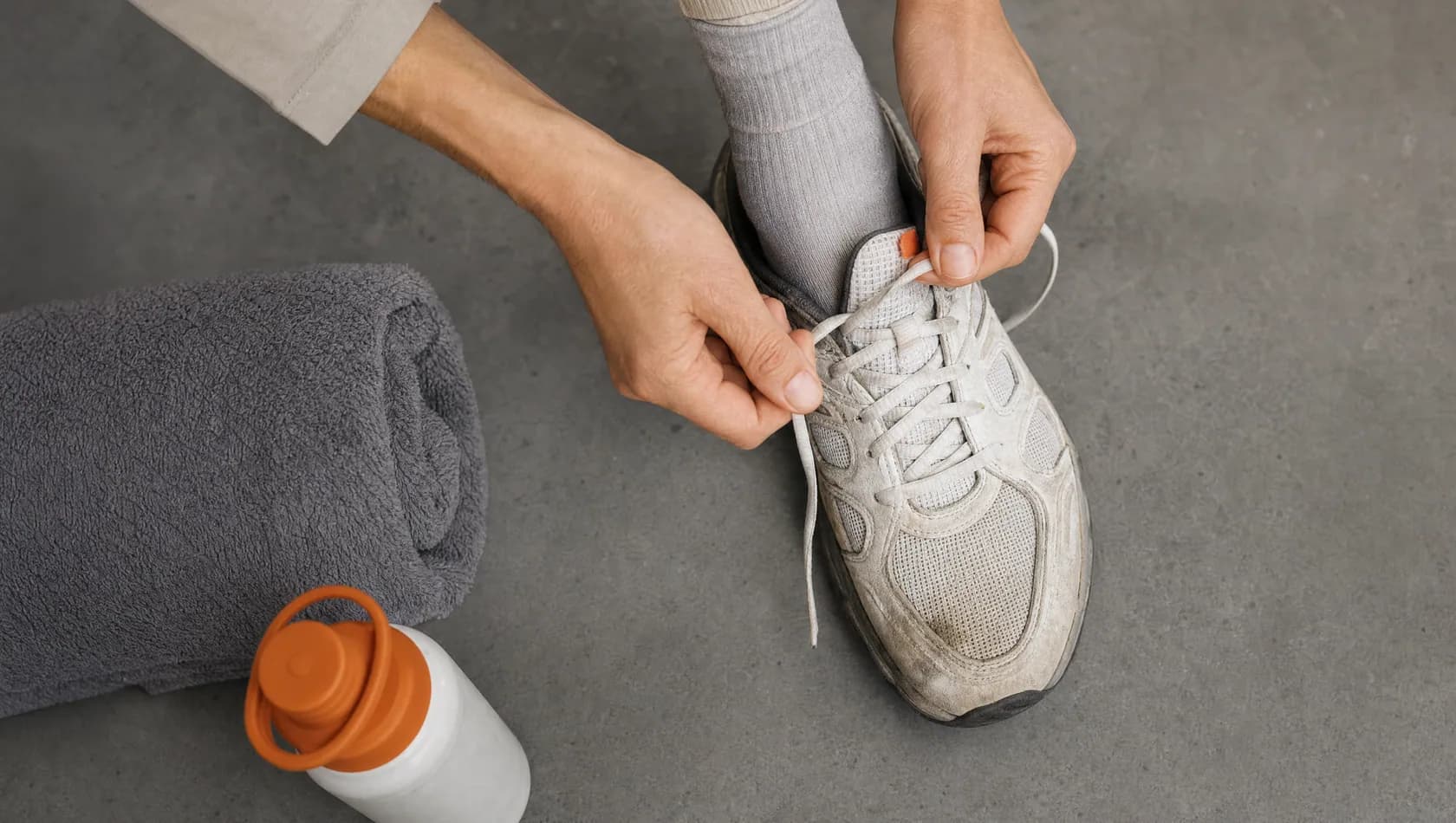 Seen from above: hands unlacing a worn trainer beside a rolled towel and a water bottle, flat even light.