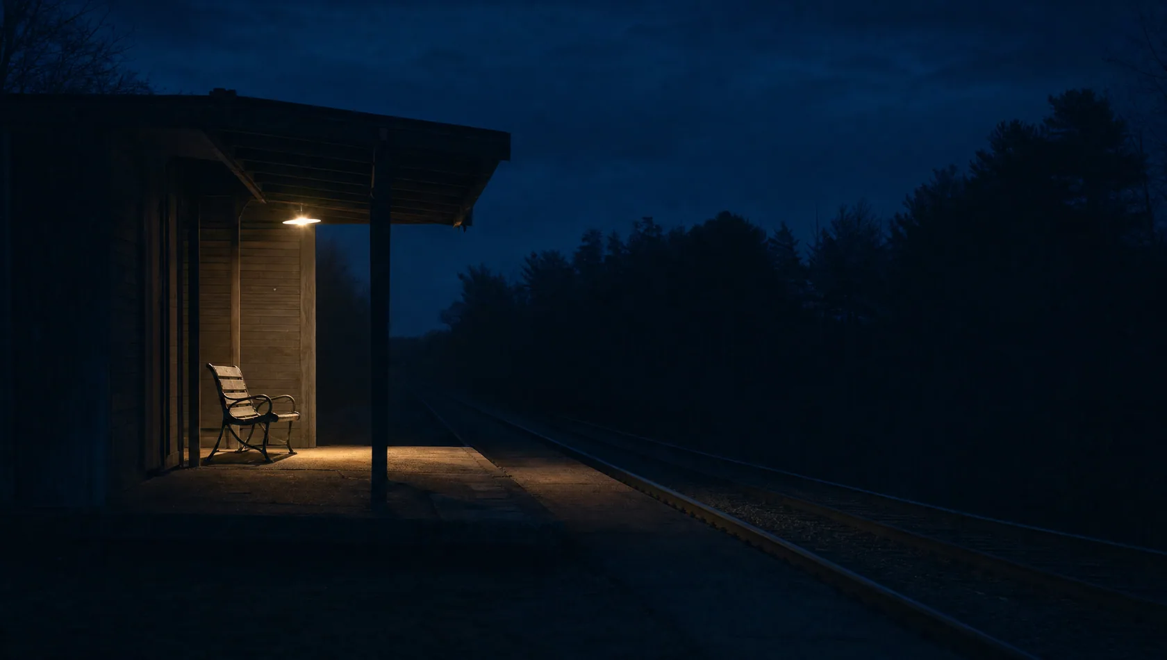 A station platform after the last train, one lamp burning over an empty bench, the track beyond in darkness.