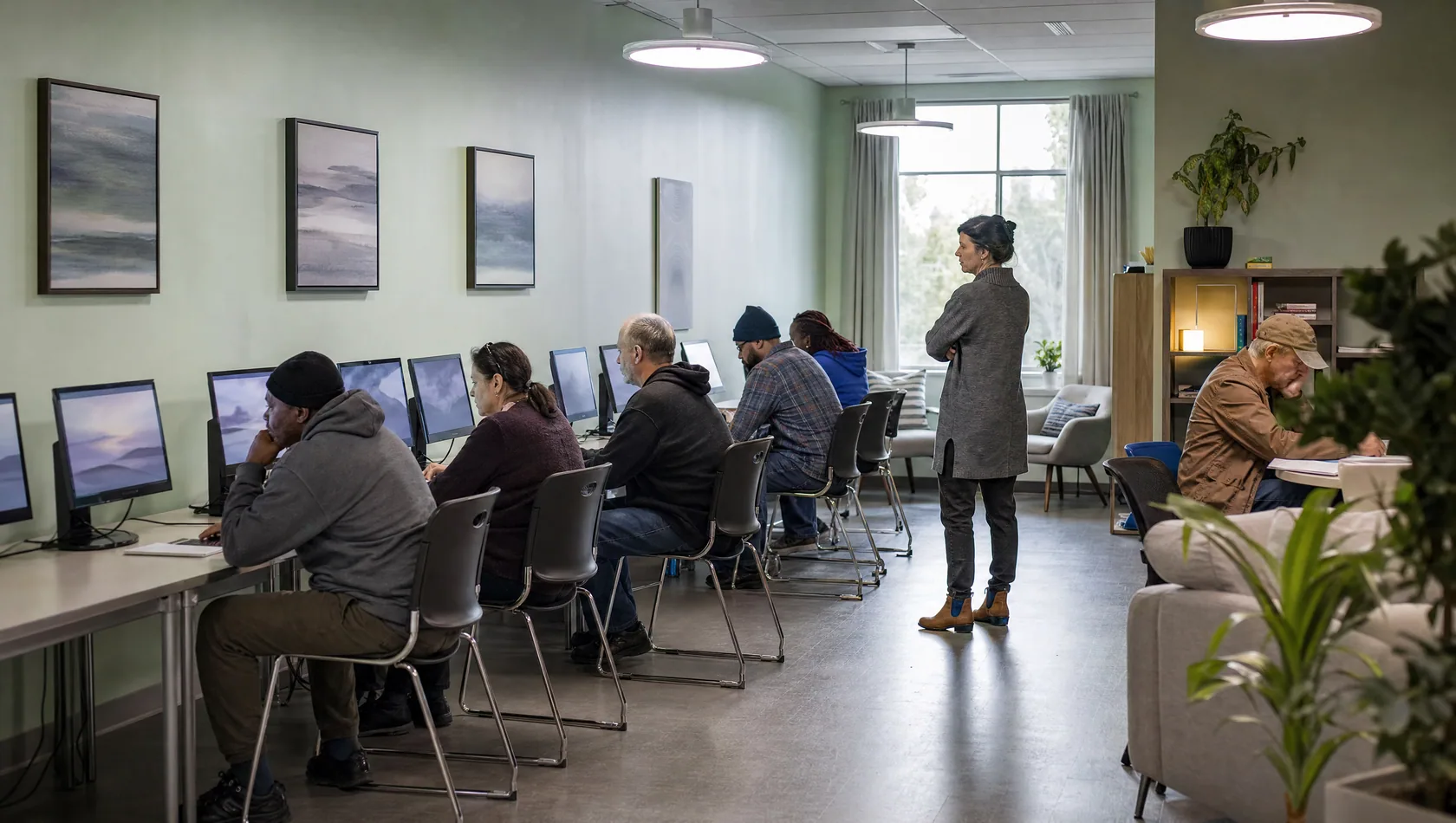 A community computer room with adults at a row of screens and a volunteer standing by, overhead light.