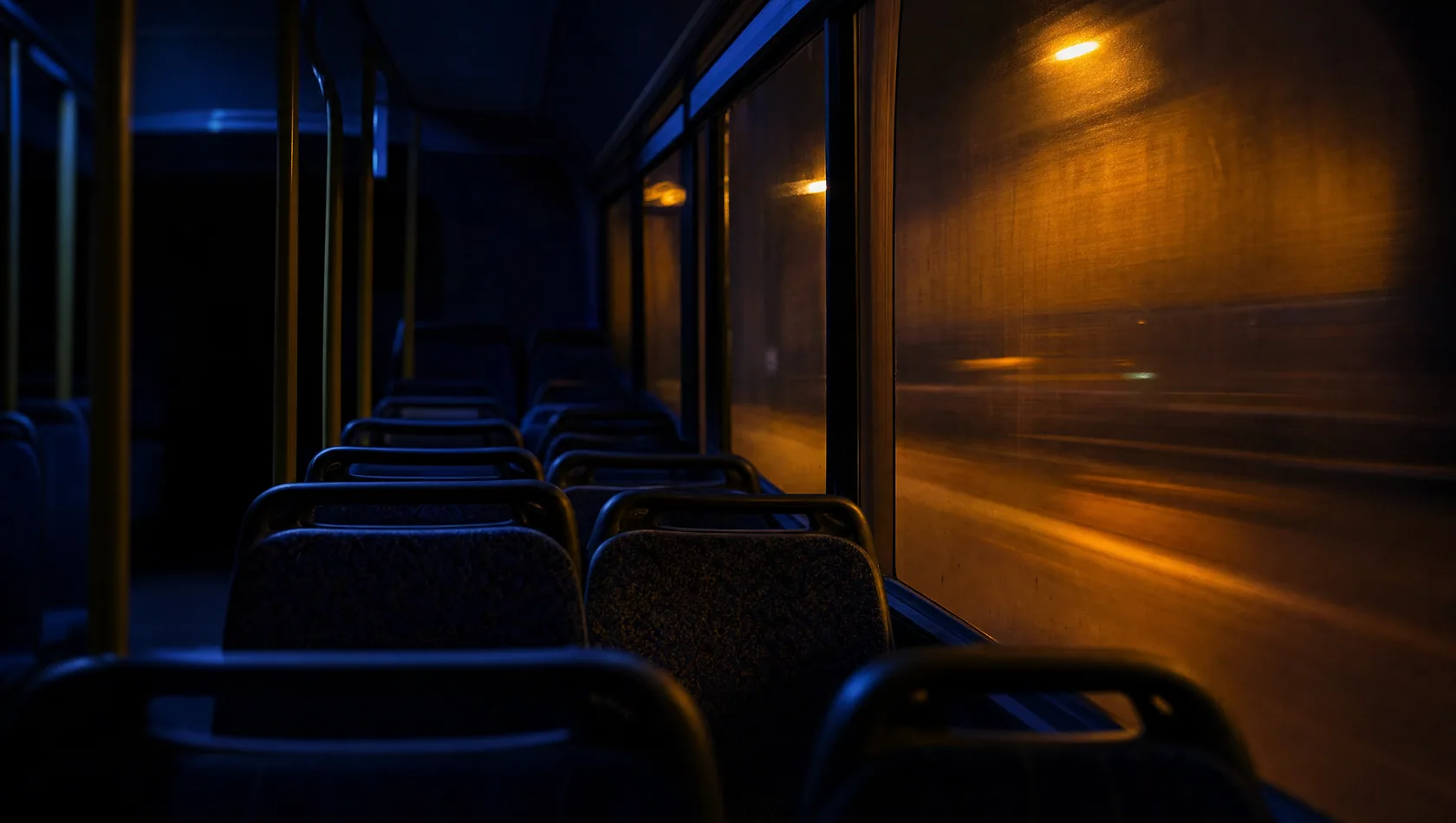 The inside of a bus at night, empty seats in rows, the window a smear of orange street light.