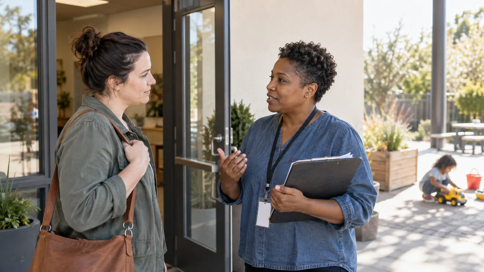 A parent and childcare worker talking at a sunny community center entrance while a child plays safely in the distant background.