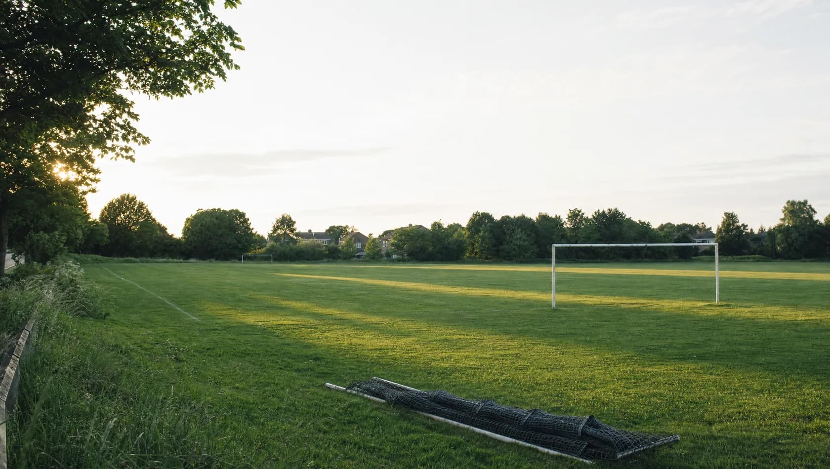 An empty suburban playing field at the golden end of the day, goalposts casting long shadows, a rolled net at the touchline.