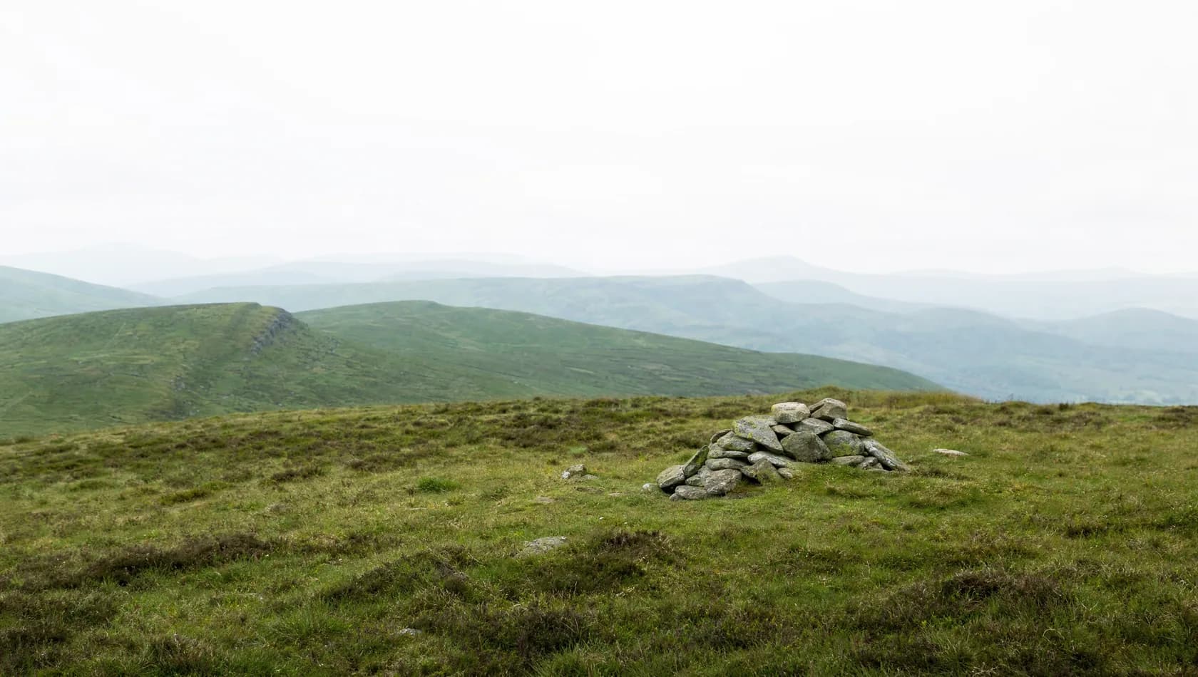 A high moorland ridge at midday with layered hills receding into haze and a low cairn of stones in the near ground, no people.