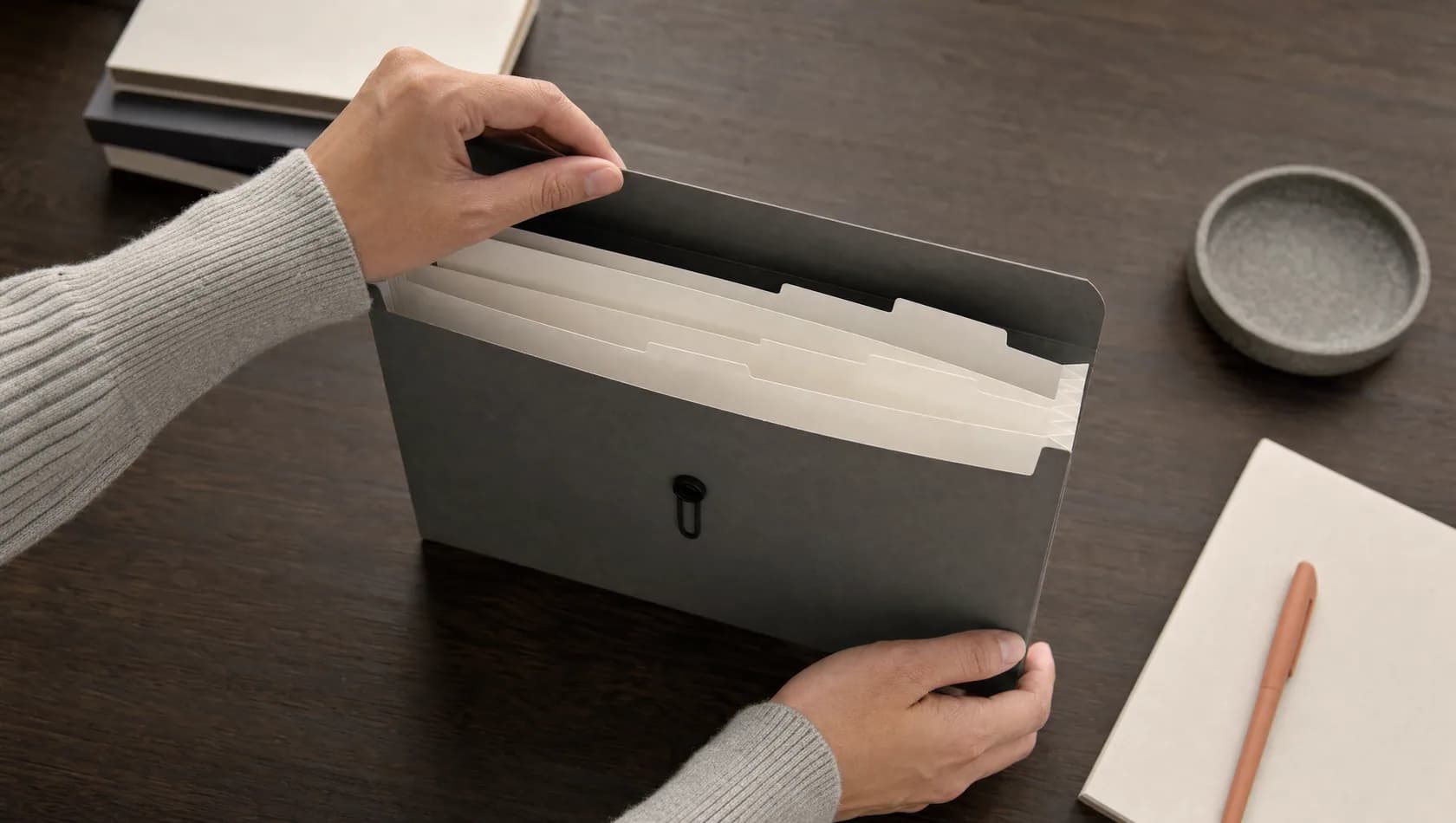 Close up: a hand lifting the flap of a concertina file with blank tabs on a dark wood surface.