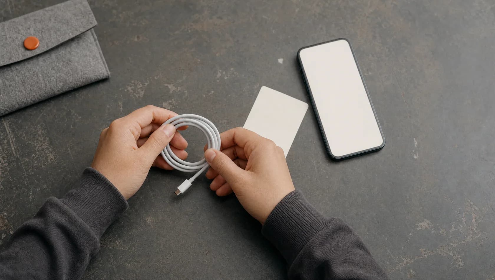 Seen from above: hands coiling a charging cable beside a plain handset and a blank card on a worn table.