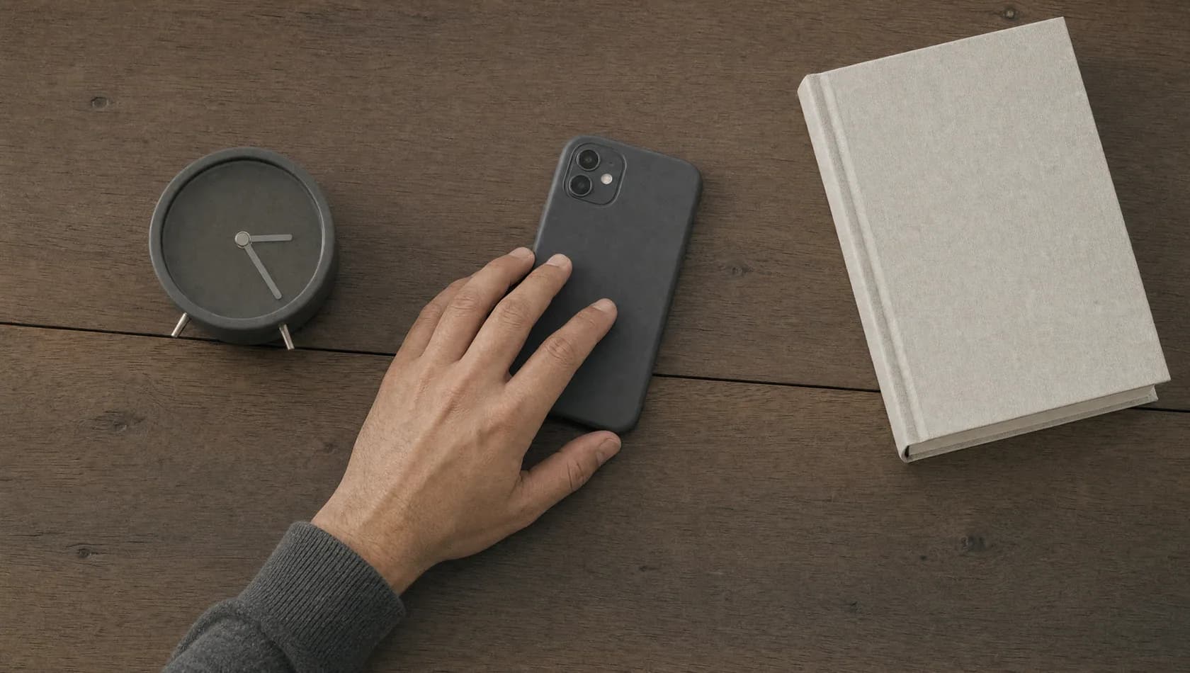 Seen from above: a hand laying a phone face down on a wooden table beside a wound alarm clock and a closed book.