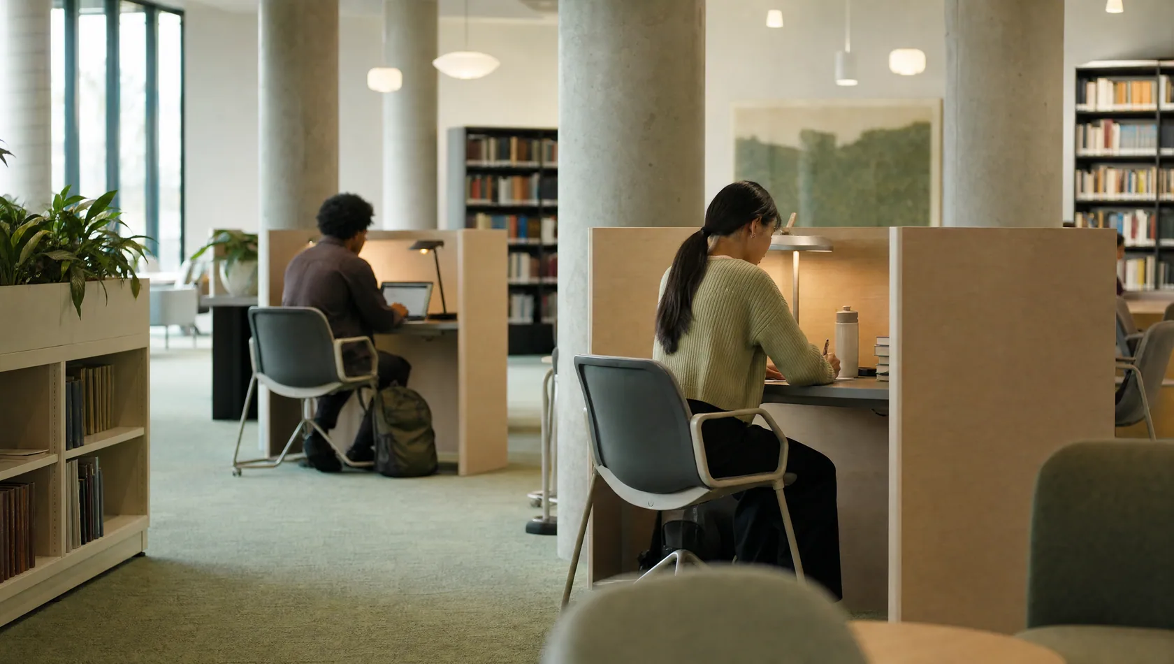 A public library study area at middle distance: partitioned desks, two people working apart, quiet daylight.