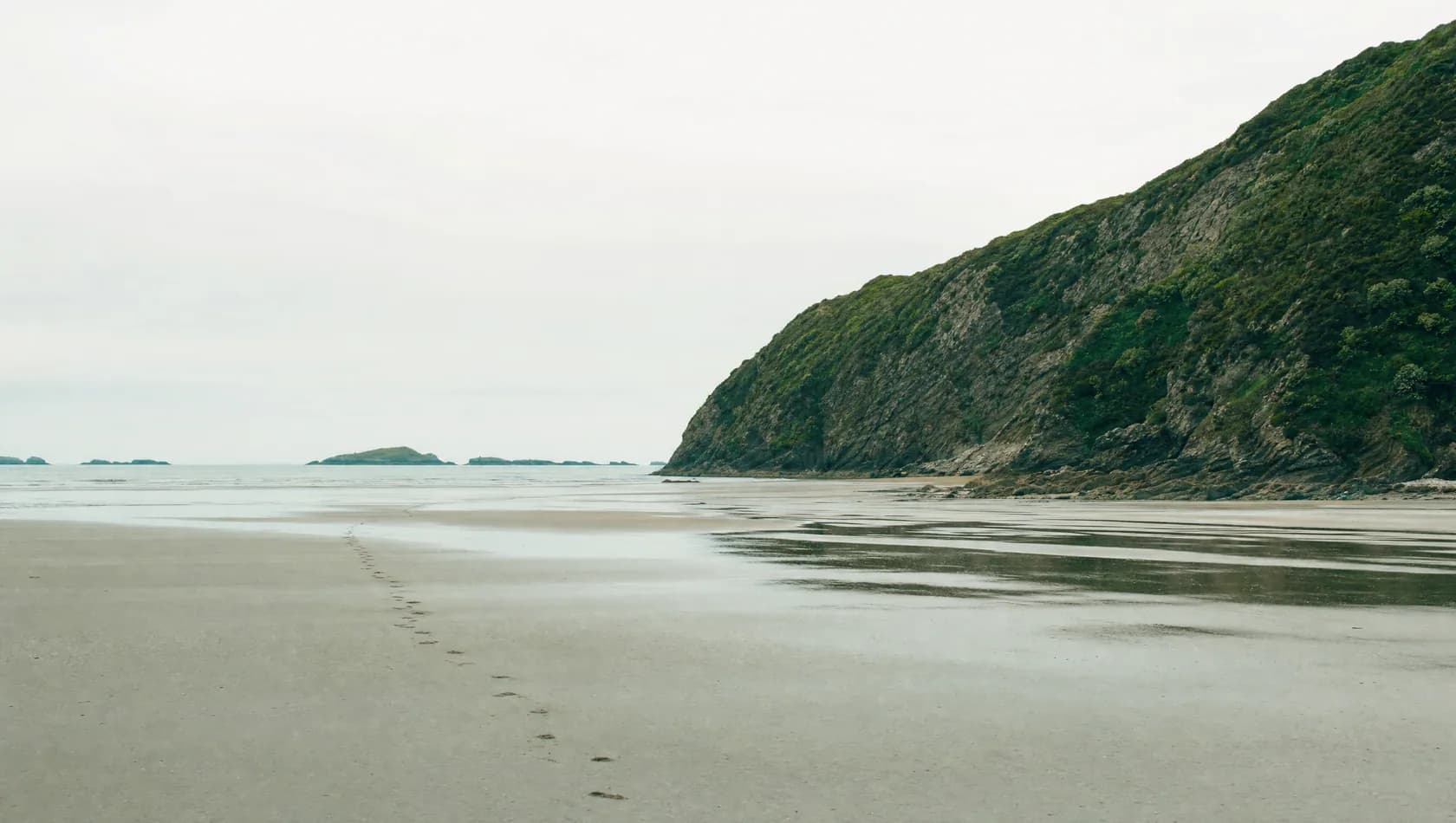 A wide stretch of open shoreline at low tide under a high pale sky, one line of footprints already softening, no people.
