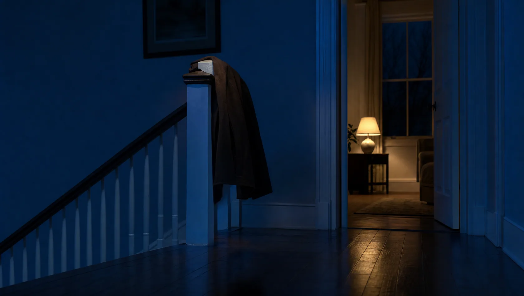 A hallway at night with a coat still on the newel post and a lamp left on in the room beyond, no people.