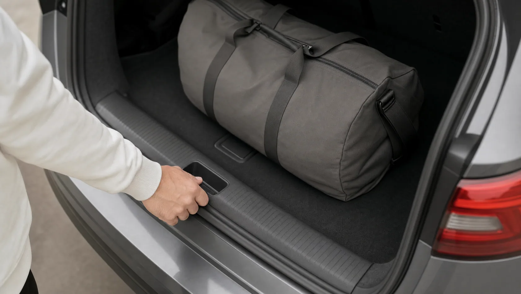Close up: a hand on the handle of an open car boot with a single holdall inside, flat grey daylight, nothing labelled.