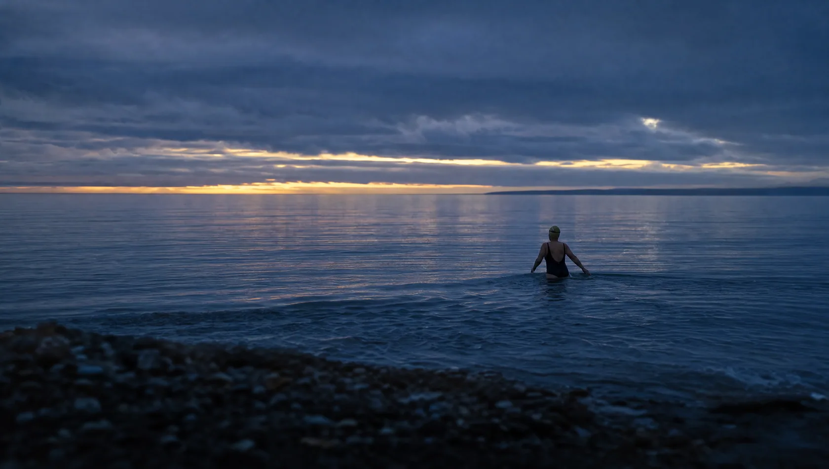 A lone swimmer wading into flat grey water at first light, seen small from the shore, cold blue with one pale gold band.