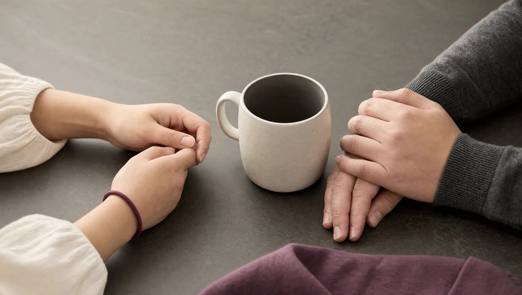 Close up: two pairs of hands resting on a table either side of a single mug, no faces, soft flat light.