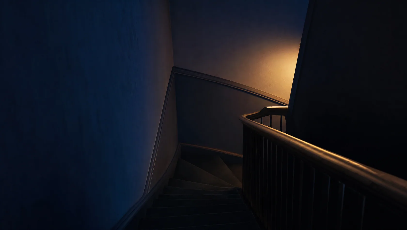 A staircase turning in half-light, the handrail catching a distant lamp and the treads lost in dark, no people.