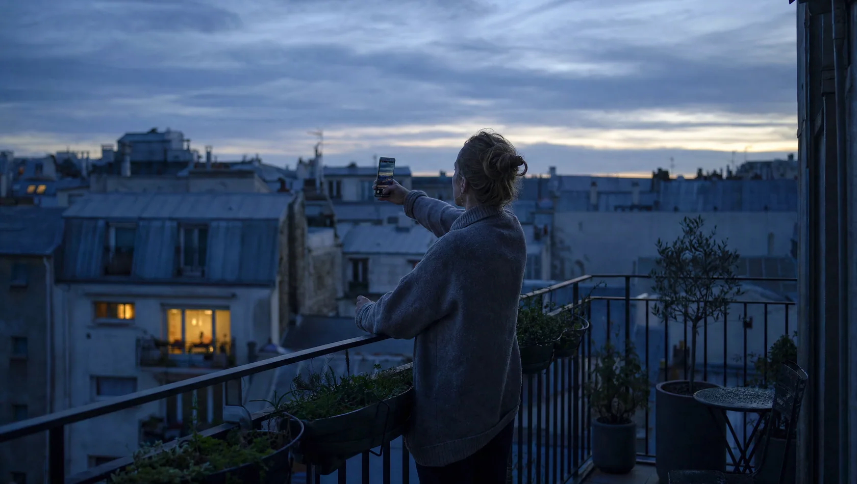 A person on a balcony at dawn holding a phone at arm's length, city rooftops behind in cold blue, one warm window opposite.