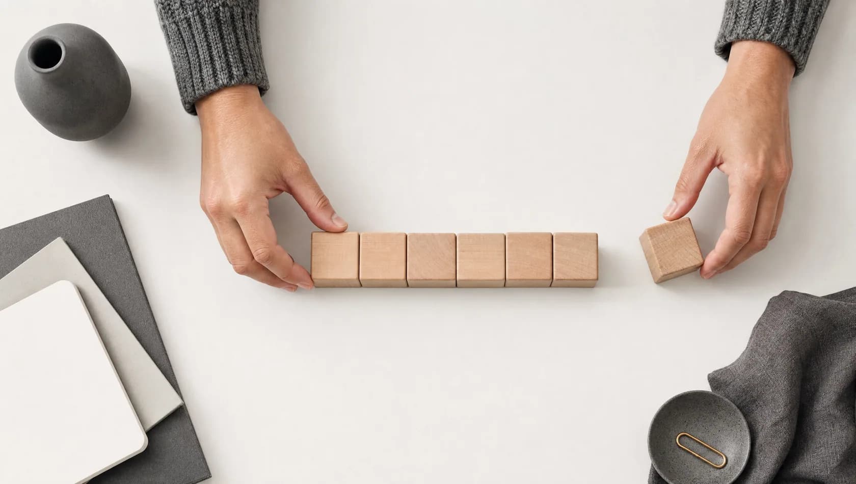 Seen from above: hands laying six plain wooden blocks in a row on a paper-white ground, one block set slightly apart.