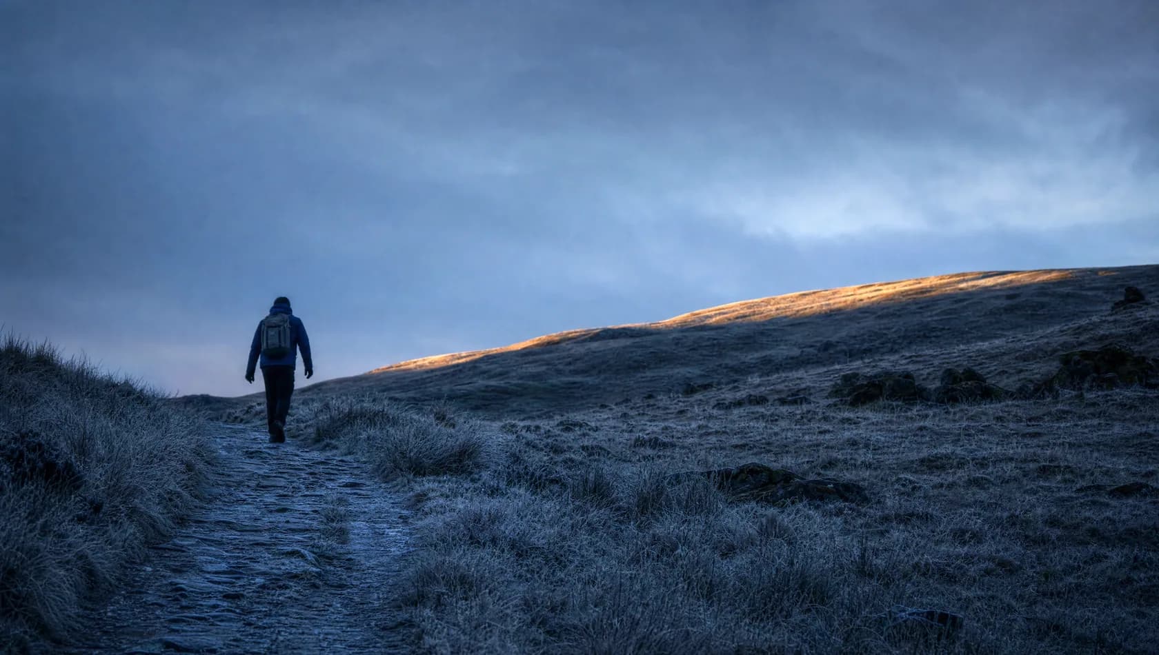 A lone walker starting up a hill track at dawn, seen small from below, frost on the grass and one warm band of light along the ridge.
