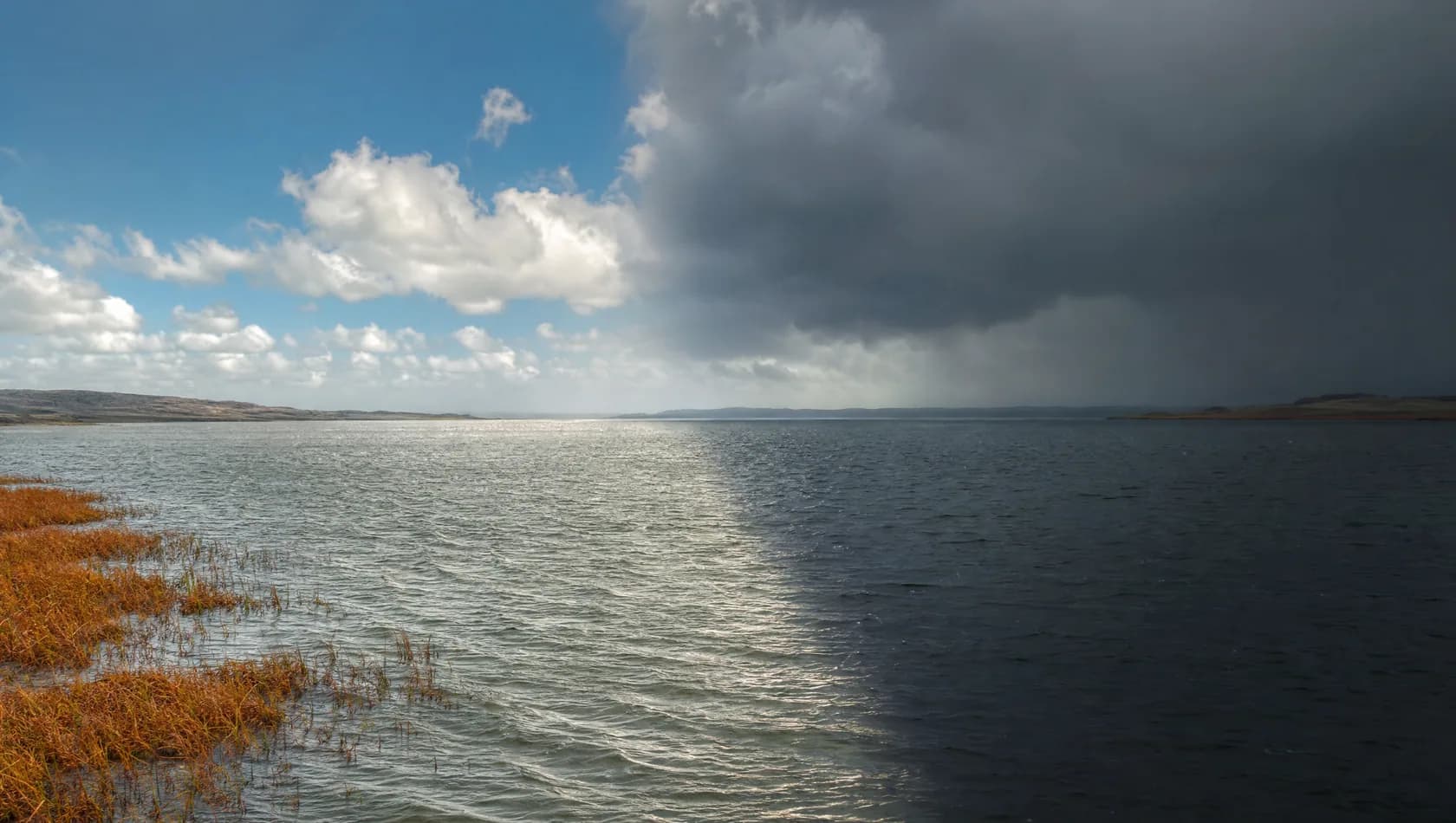 A bright squall crossing a wide estuary, one half of the sky clear and the other slate grey, the water divided between them, no people.